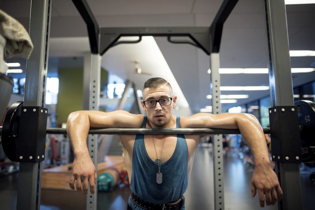 A student wearing a tank top and dog tags rests in an exercise rack during a workout.