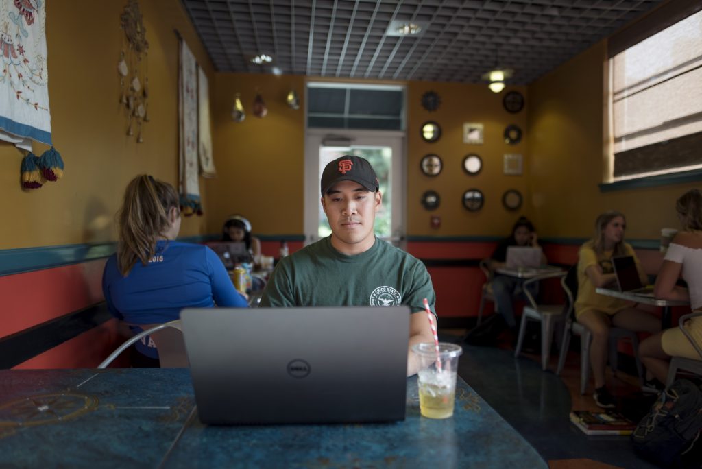 A seated student wearing a baseball cap works on a laptop in Creekside Coffee.