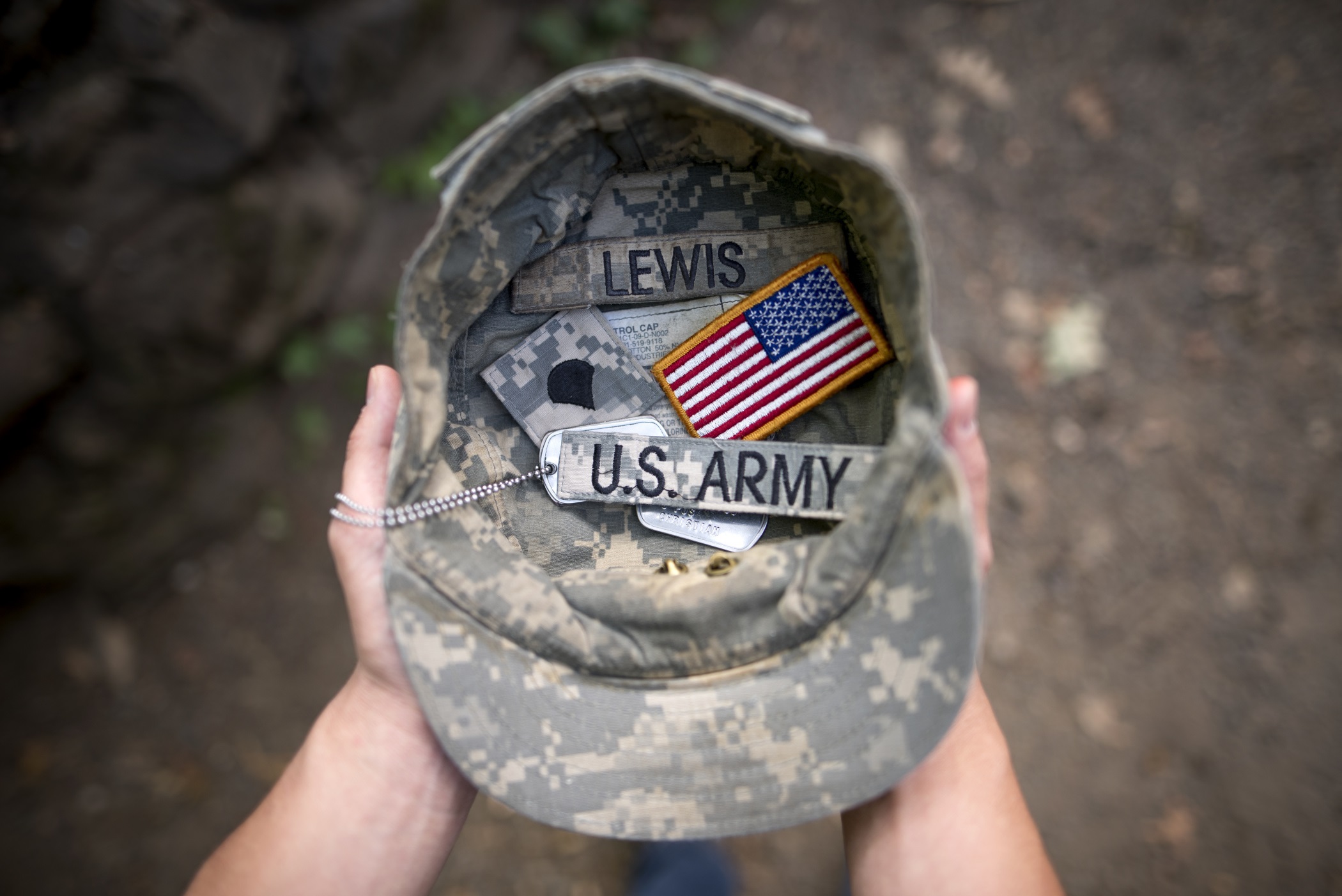 Two hands hold an Army cap, which contains two dog tags, an American flag patch, and tags reading "Lewis" and "U.S. Army."