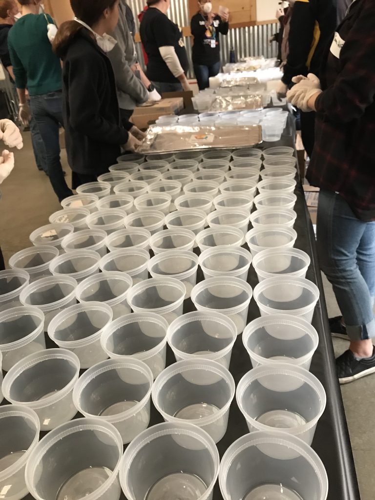 Dozens of empty soup containers are lined on a table surrounded by volunteers who will soon fill the containers with piping hot tomato soup.