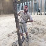Portrait of Matthew Day in his zoo keeper uniform posing next to a tree branch stuck in the sand.