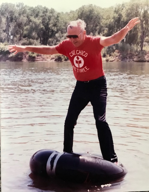 John Westlund stands and balances on an inner tube on the river while wearing pants and a shirt that says "CSU, Chico Retiree"