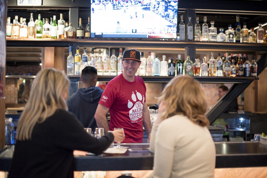 Josh Leavy smiles from behind the bar while wearing a Chico Make a Difference Day shirt.