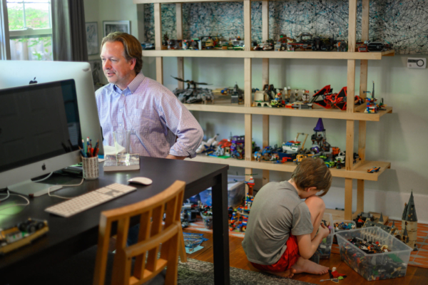 A faculty member addresses his class on his laptop, while his son assembles Legos on the floor behind him.