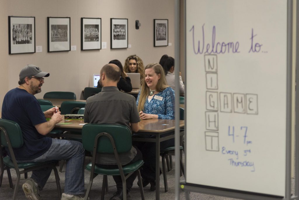 A group of people sit at a table playing board games in the library. A white board sits next to the table and reads "Welcome to Game Night."