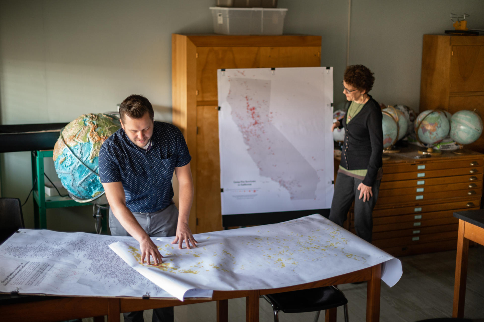 Peter Hansen and Jacque Chase look at maps on a table and on a cabinet.