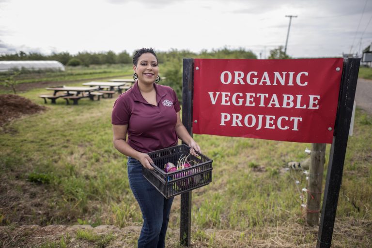 Kaeli McCarther stands next to a sign that reads "Organic Vegetable Project" while holding a crate of vegetables.