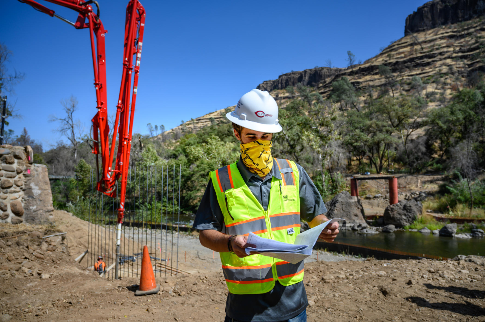 A student in a hard hat looks at engineering plans as heavy equipment is used in a bridge rebuild in a canyon behind him.