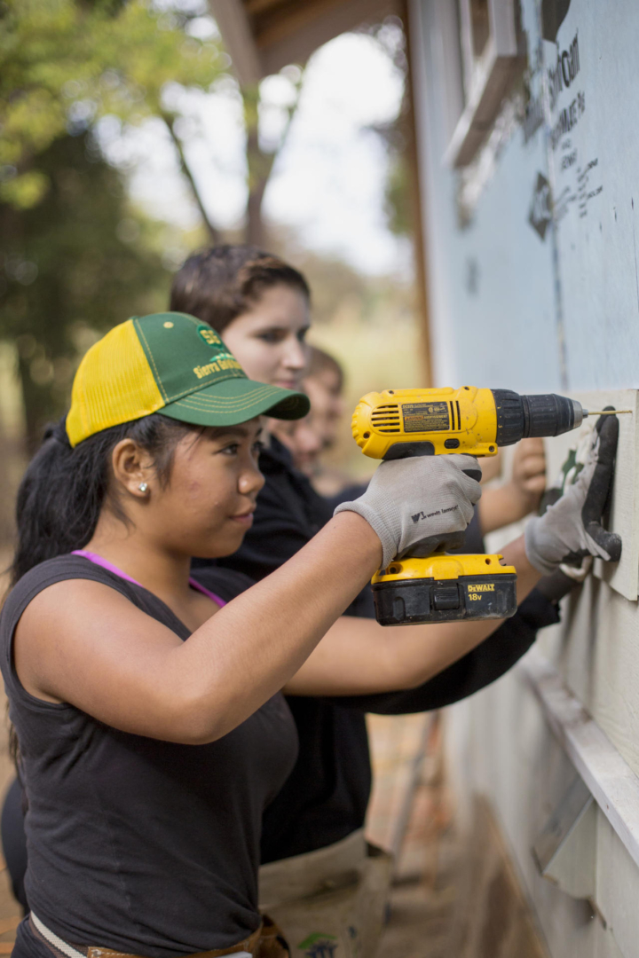 A student uses a battery-powered drill to install siding on a house.