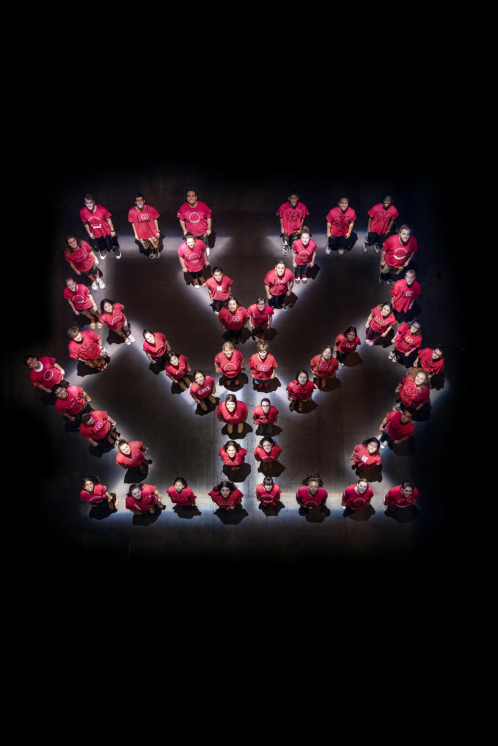 Students in red T-shirts look skyward while standing in a pattern of interconnected arrows, the symbol for CAVE.