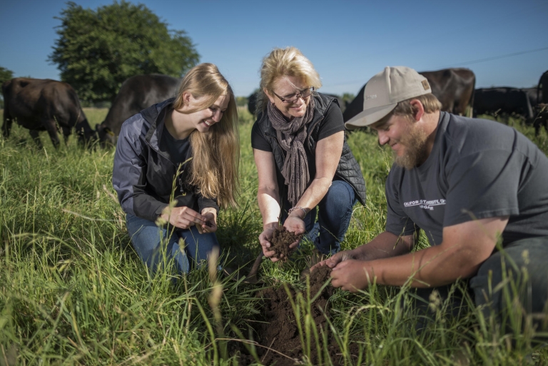 Cindy Daley and two students crouch in a field of grass holding piles of soil in their hands.