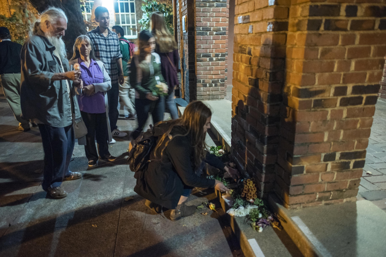 A group of several people somberly pay respects with notes and flowers in front of a brick pillar by Kendall Hall.