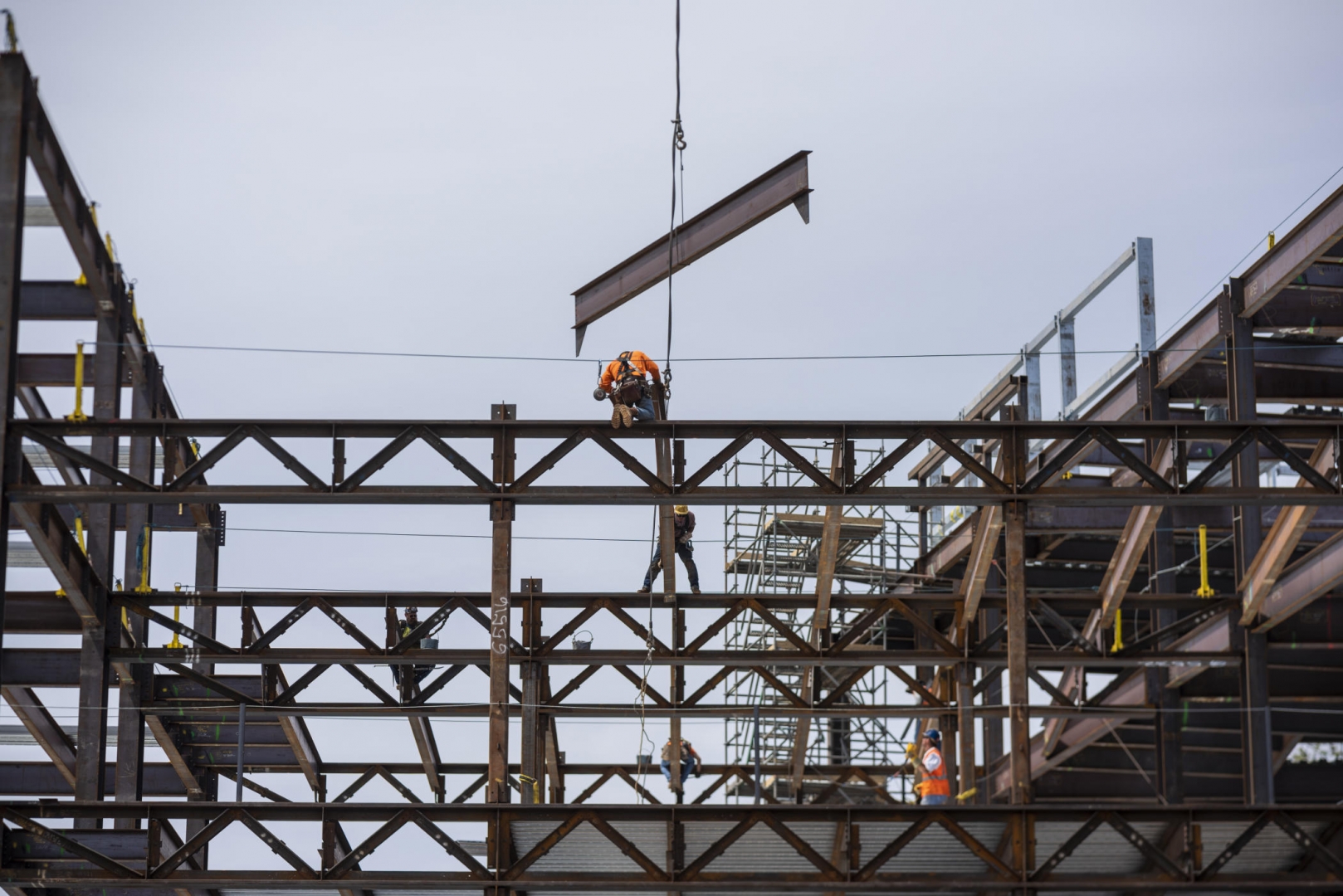 A cable moves a large steel beam over scaffolding and a steel frame as construction workers prepare to put it in place.
