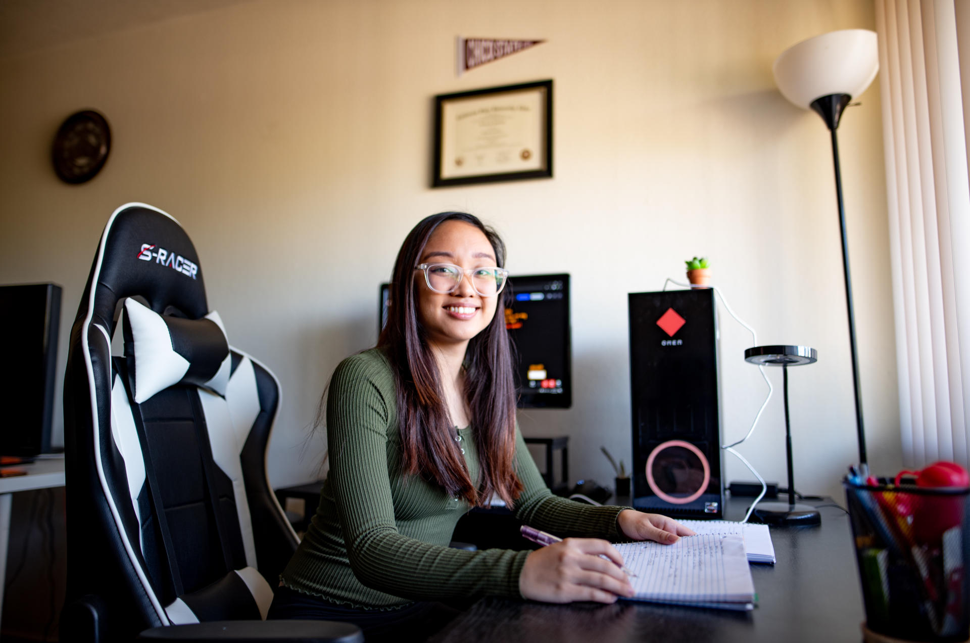 Portrait of a student seated at a desk in her home, surrounded by electronic equipment.