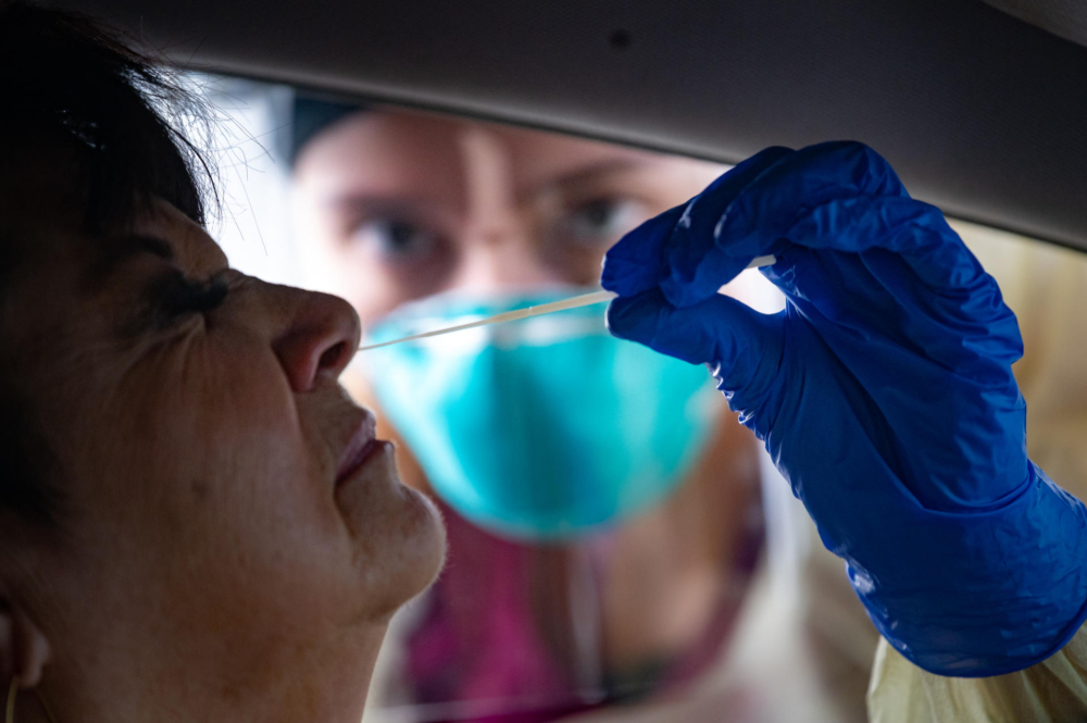 A nursing student wearing gloves administers a nasal swab test to someone sitting in a car.
