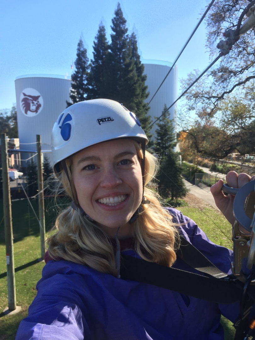 Wearing a helmet and big smile, Cheyenne Arrington hangs from the tops of the ropes course with the Wildcat logo visible on the boiler-chiller tower behind her.