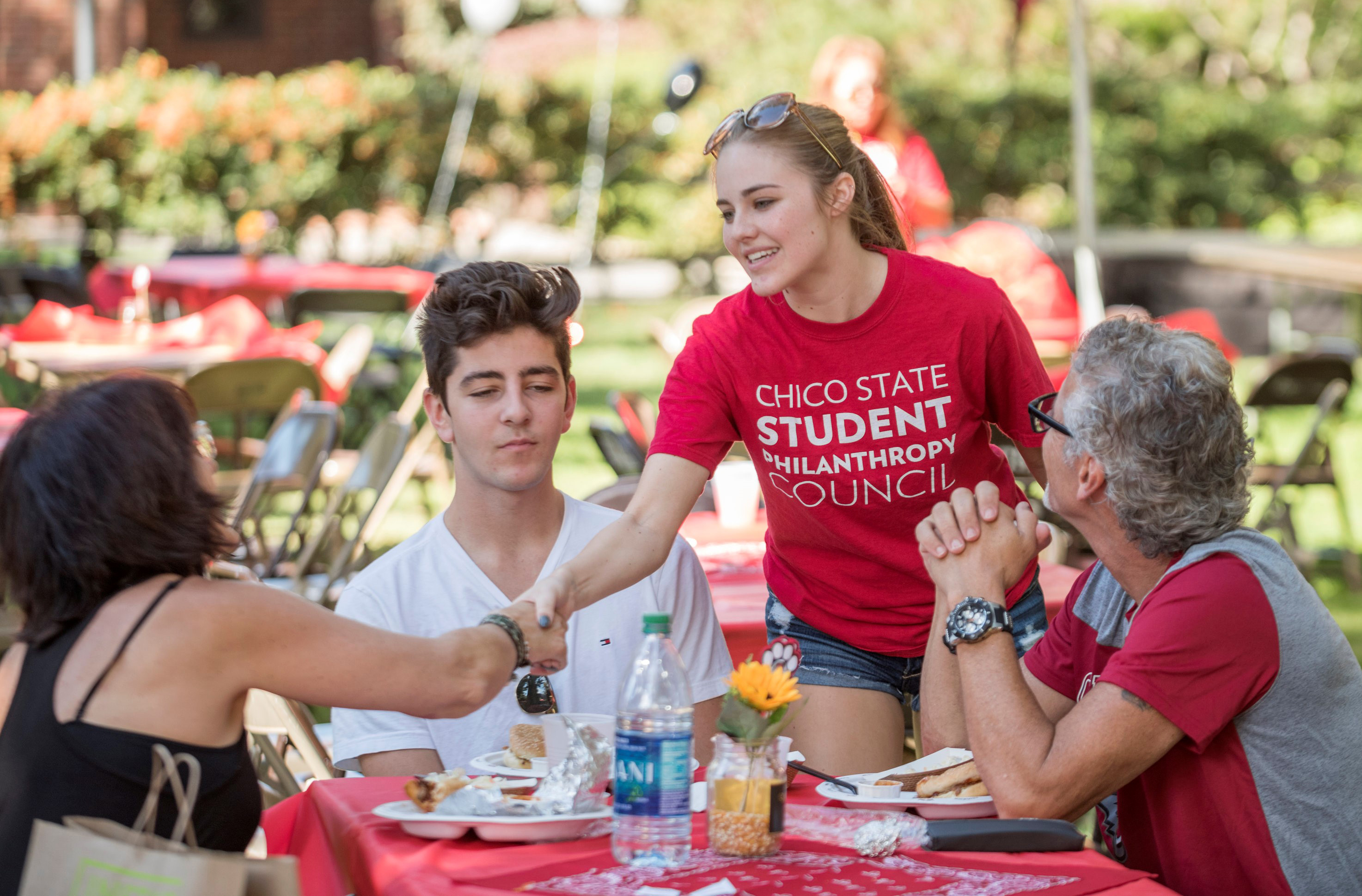 Elizabeth, in a Student Philanthropy Council t-shirt, brightly greets a student and their parents at the Family Weekend BBQ