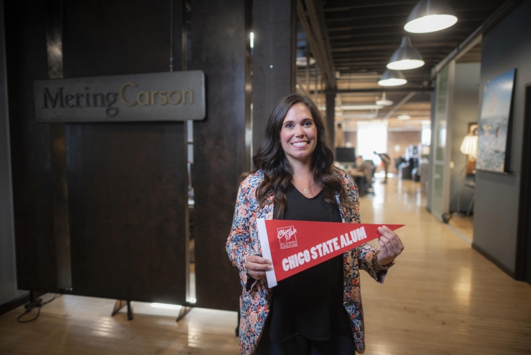 Michelle McIntosh smiles and holds a Chico State alum pennant at her Mering Carson office.