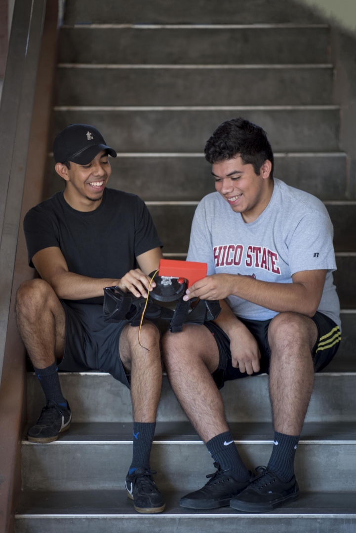 Two students sit on a stair, smiling and together holding the robotic knee brace prototype they built.