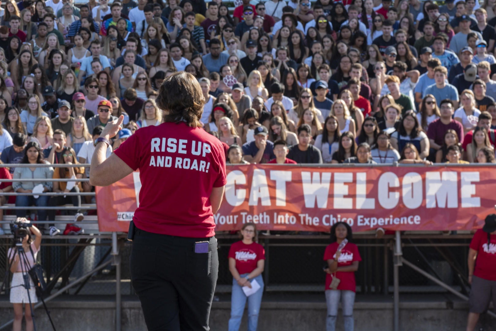 President Gayle Hutchinson speaks to a crowd of students filling bleachers at University Stadium with the words "rise up and roar" visible on her back.