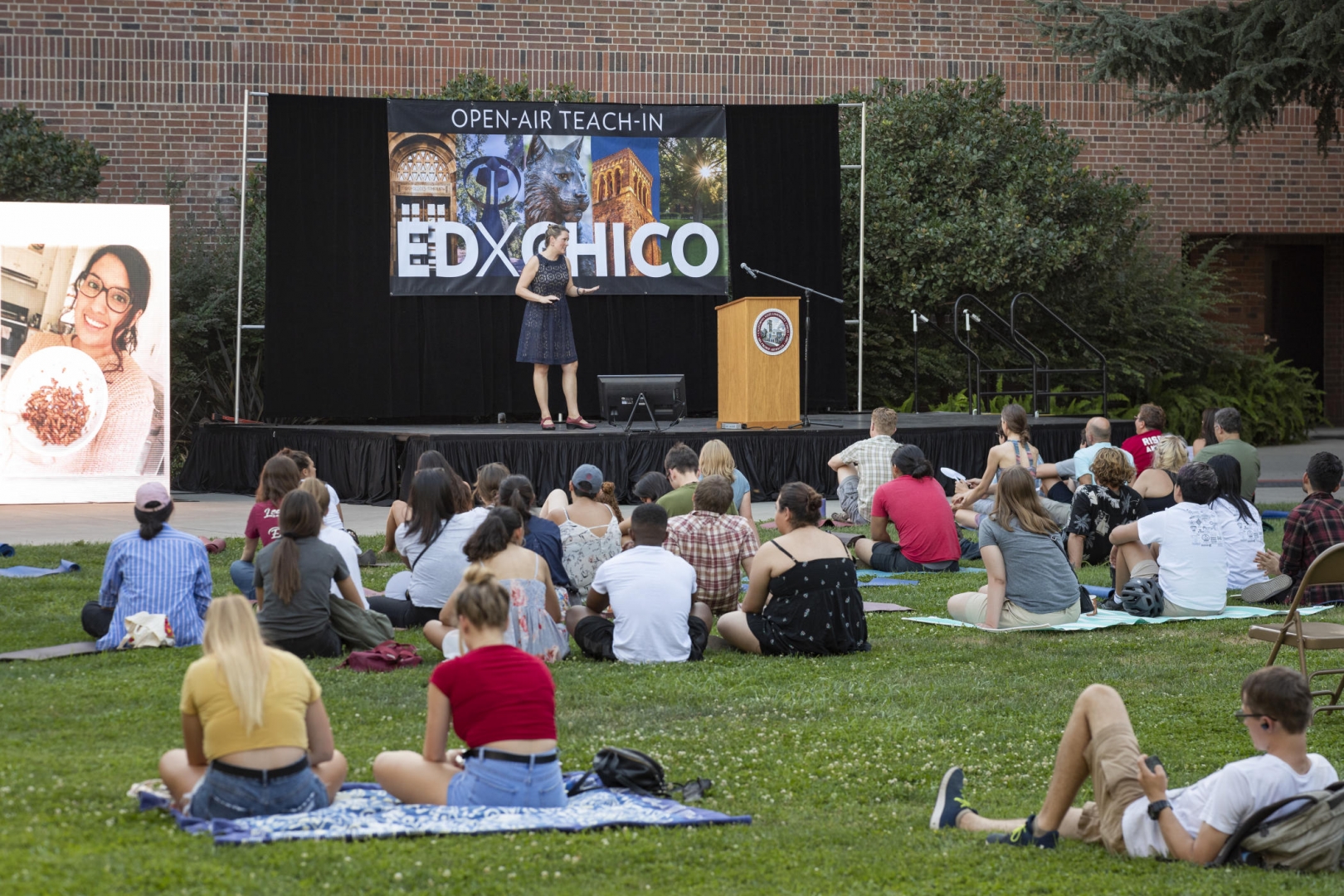 Students sit on a lawn watching a presentation by faculty on a stage in front of a screen that reads E-D-X Chico.