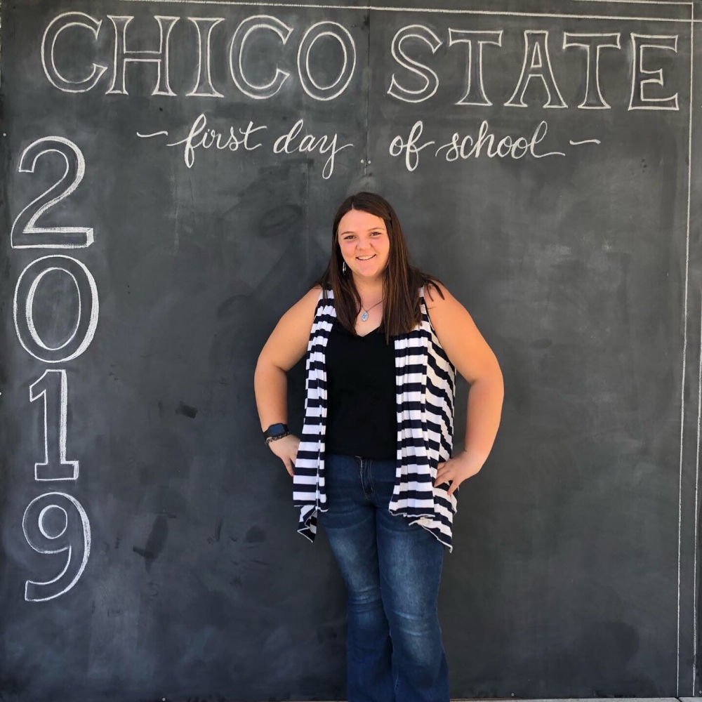 Stephanie Mills stands in front of a chalkboard sign that says "Chico State First Day of School 2019."