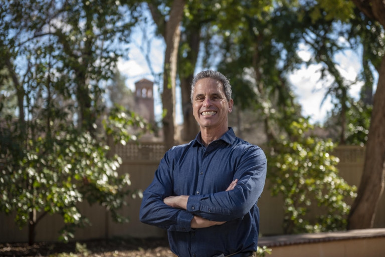 Darryl Schoen smiles with his arms folded in front of trees and Trinity Hall.