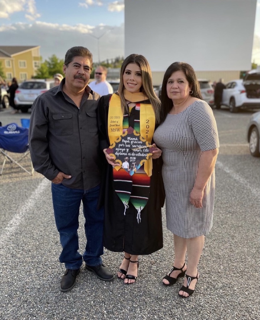 Veronica Ledesma stands between her parents, while dressed in a graduation gown and holding a graduation cap.
