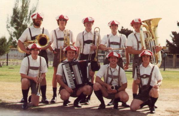 Nine men stand holding their various musical instruments outside while wearing lederhosen and red and white hats.