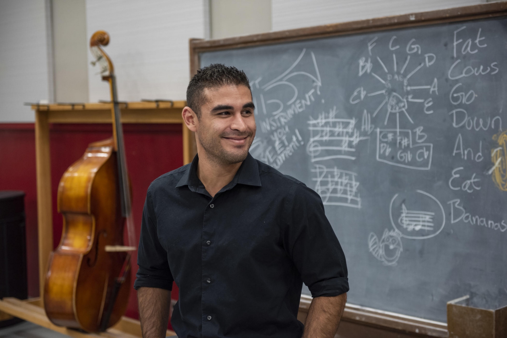 Chris Navarrete smiles and stands in front of a blackboard and a cello