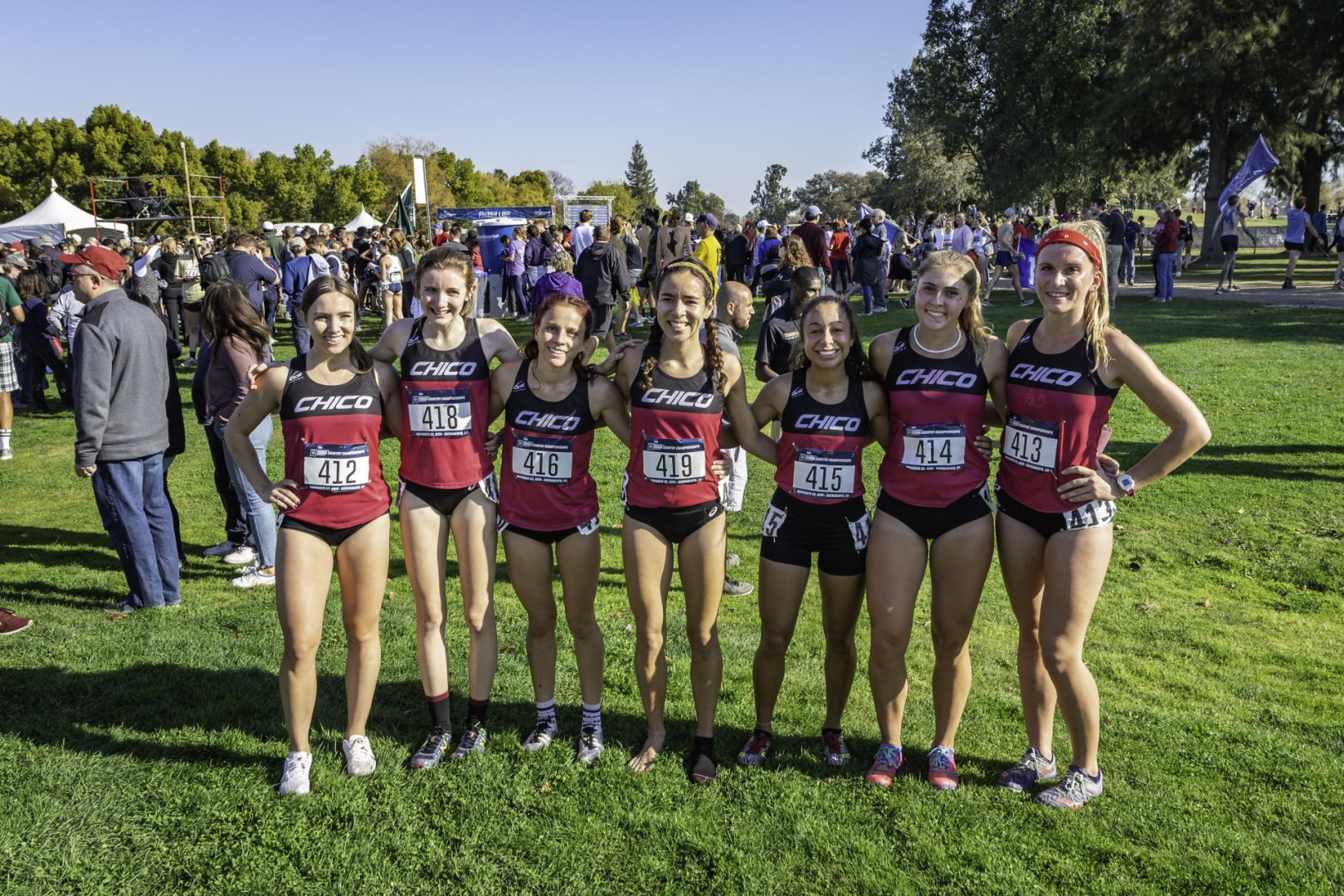 A women's cross country team poses after a race, with a crowd of people behind them.