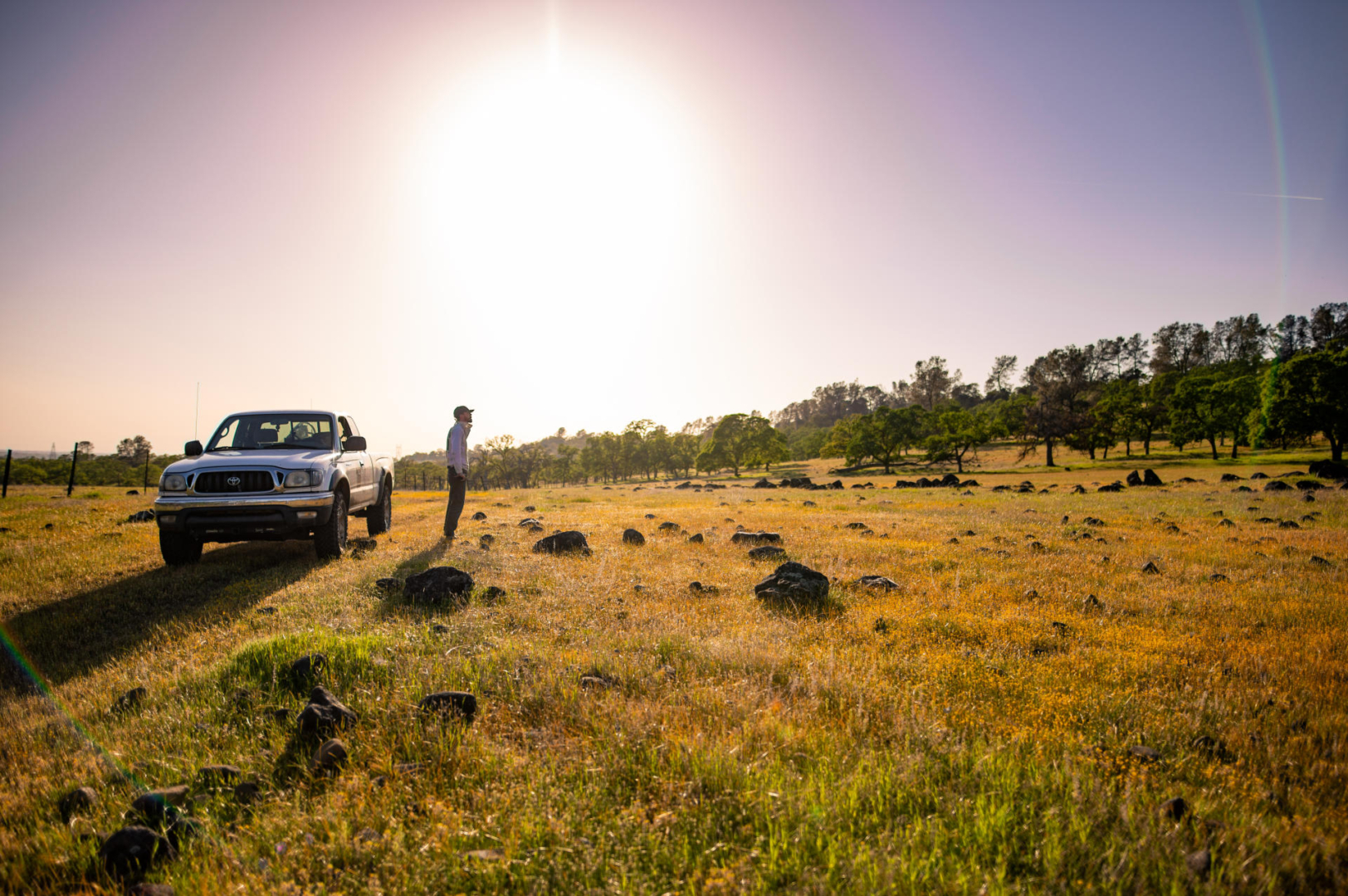 Eli Goodsell stands to the side of a truck among rocks jutting out of the grassy ground, with trees in the background and the sun beginning to set.