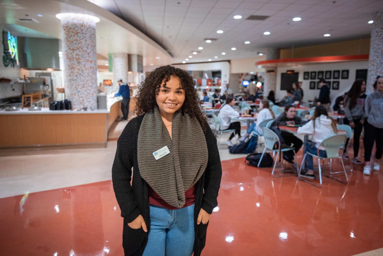 Jessica is wearing a scarf and is smiling and standing inside a dining hall with other students sitting and eating around her.