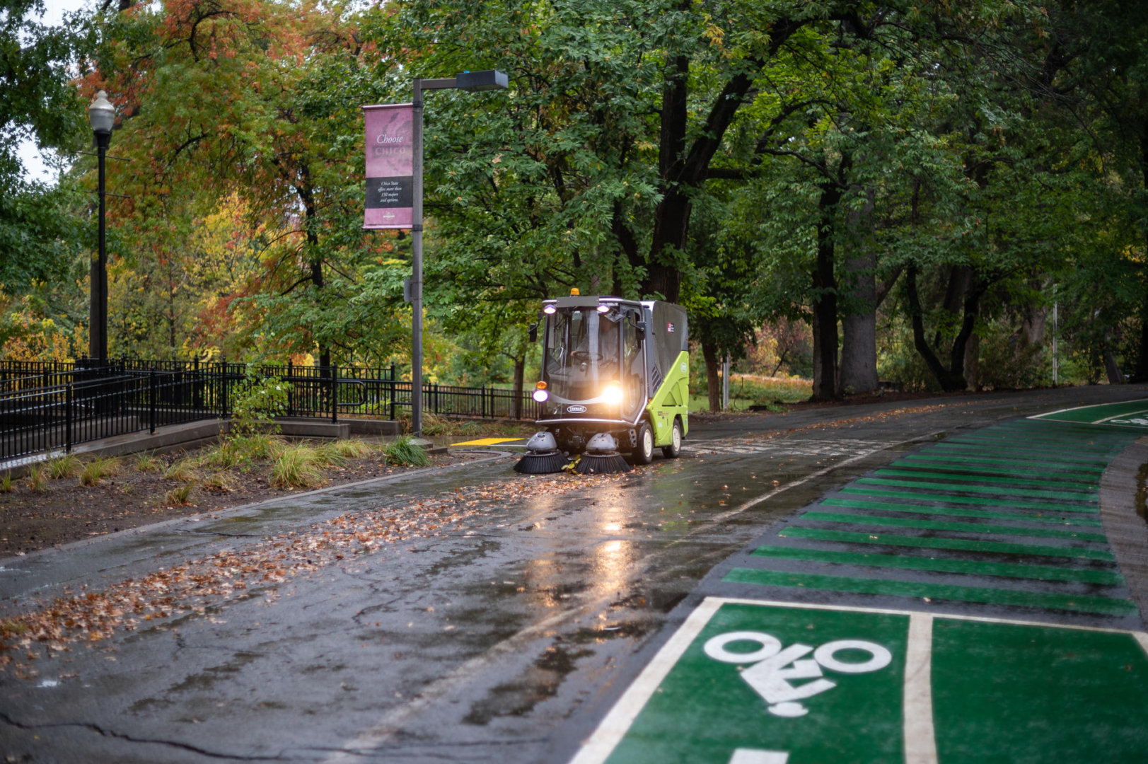 An all-electric street sweeper picks up leaves on a rainy road.