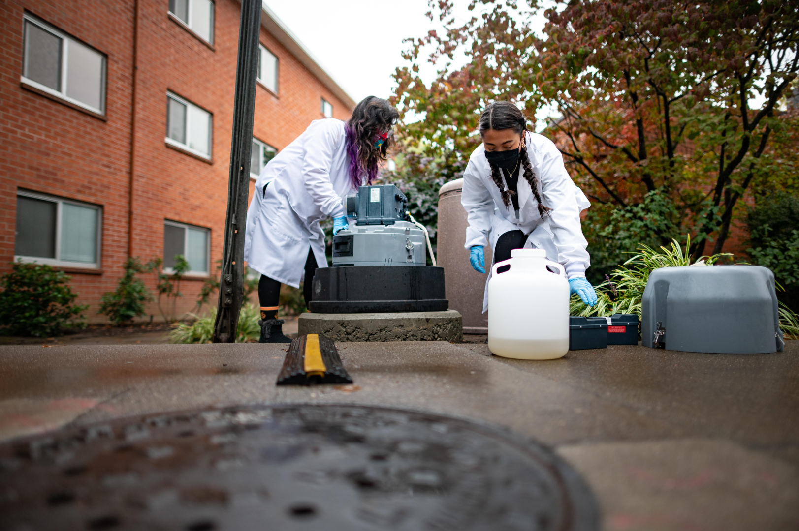 Two students wearing white lab coats collect wastewater at a special station installed outside a residence hall.
