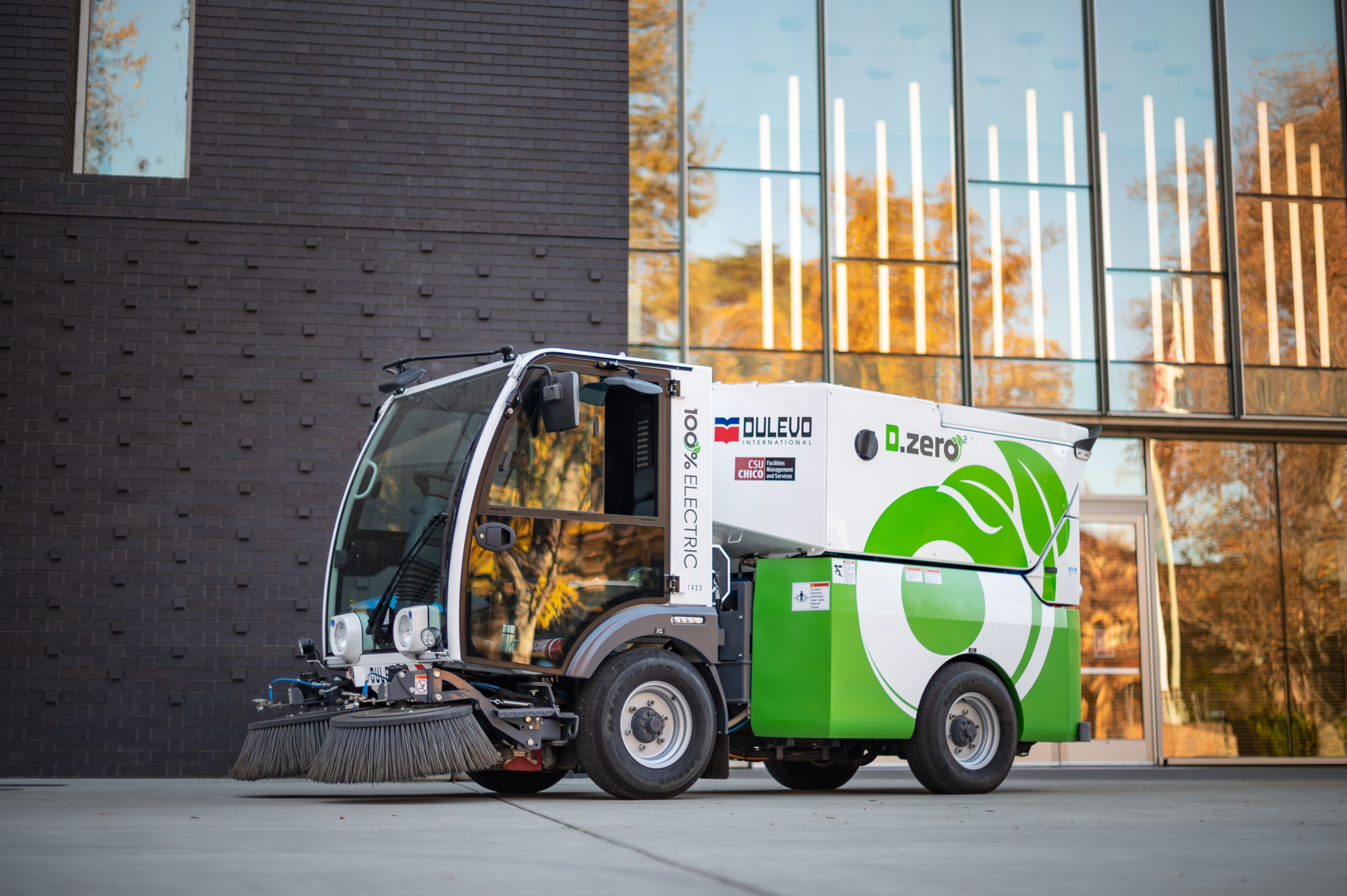 An all-electric street sweeper is parked outside an academic building.
