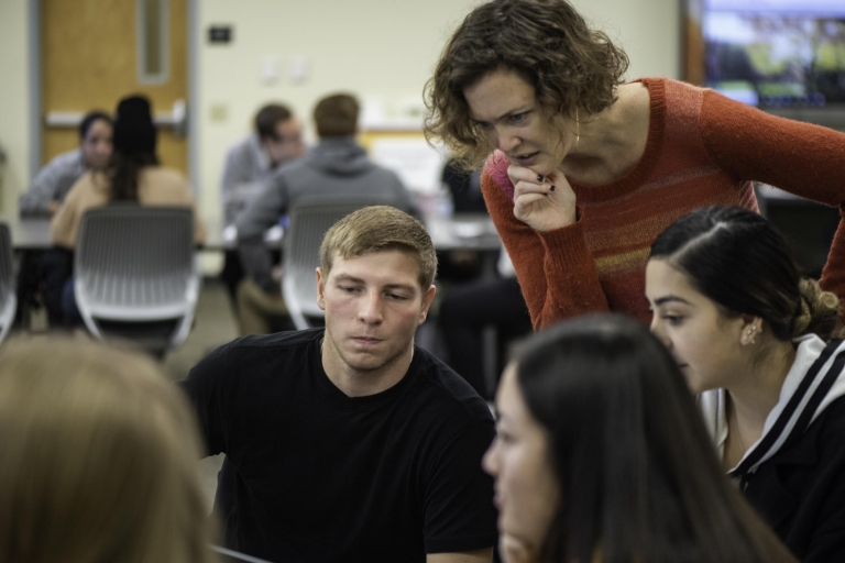 A faculty member leans over a group of students to help them work through a problem.