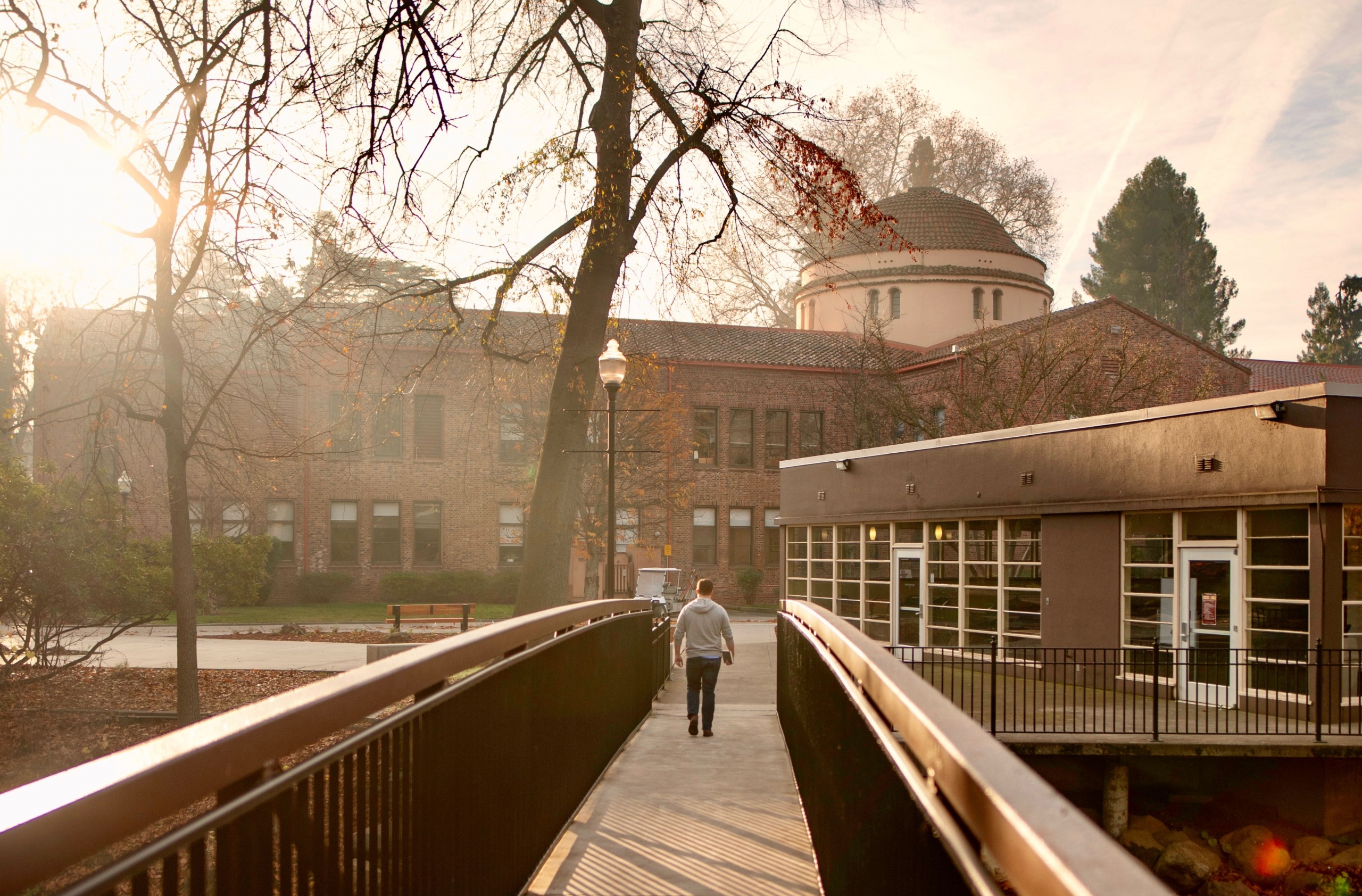 A person walks across a campus bridge, toward the rising sun.