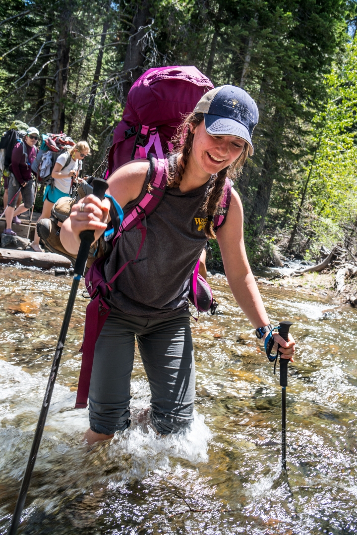 A student uses hiking poles as she walks through a knee-high creek with her backpacking pack on. 