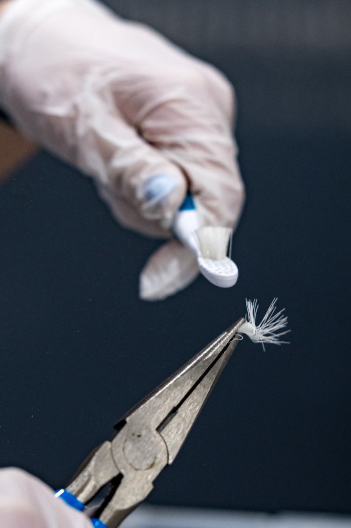 Needle-nose pliers hold a clump of toothbrush bristles while a nearby hand holds out a toothbrush they were plucked from.