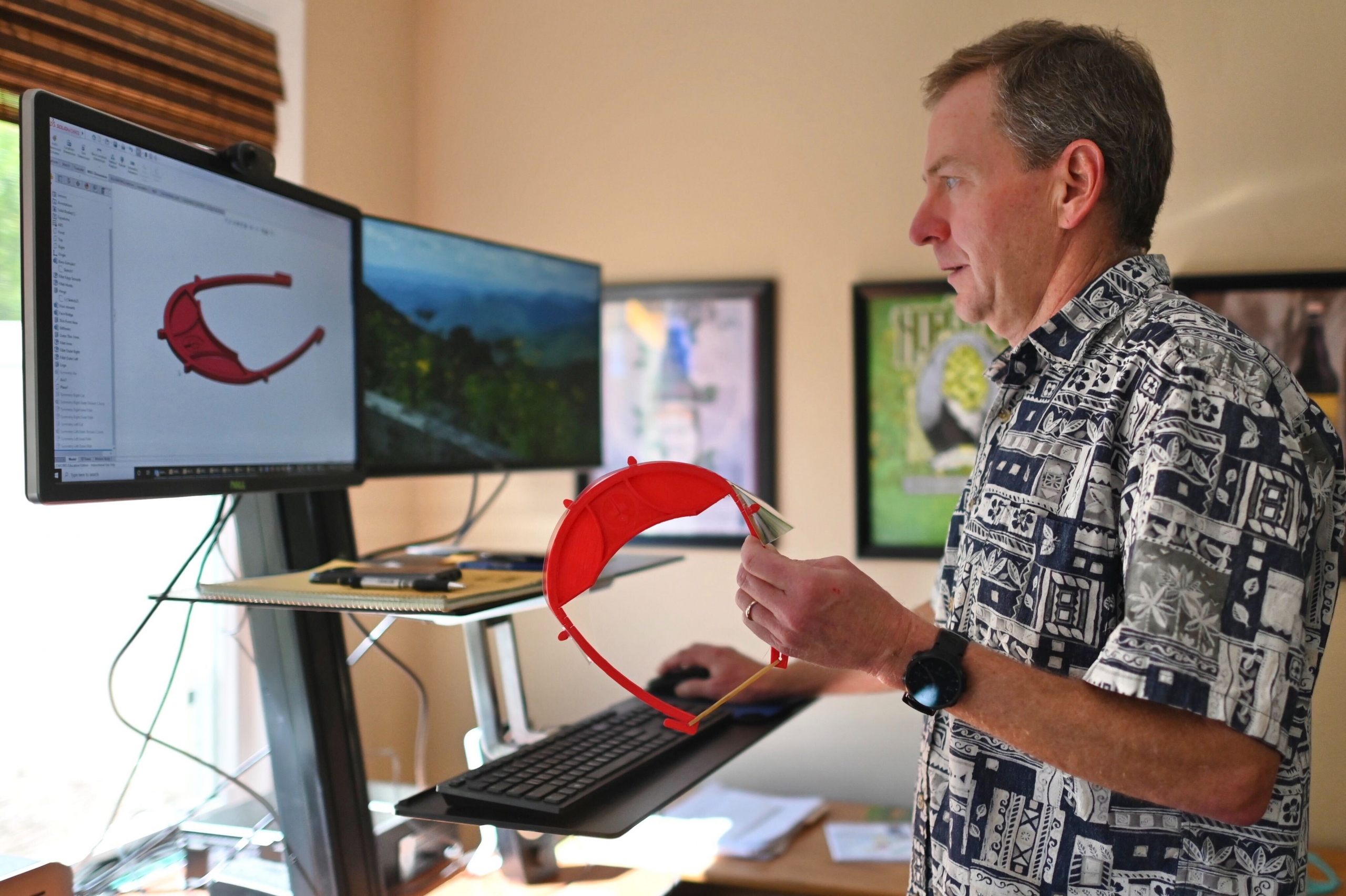 Greg Watkins stands at his computer holding a face shield frame that was created by a 3D printer.