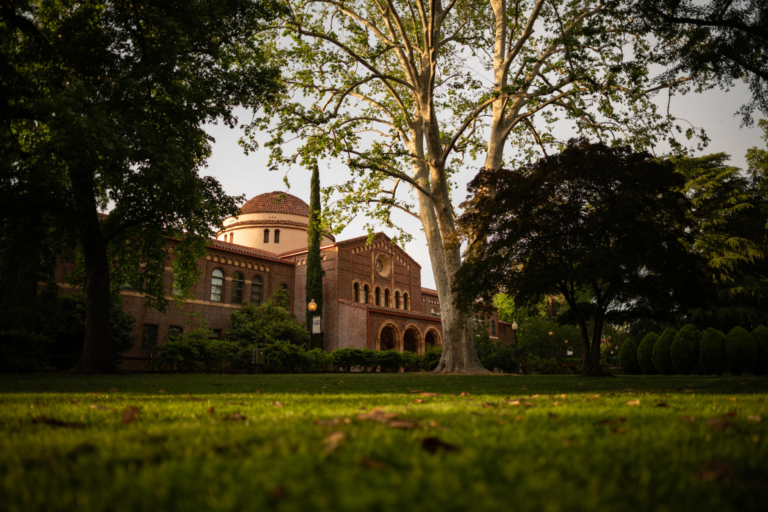 An academic building sits amongst majestic trees with a lush green lawn in front.