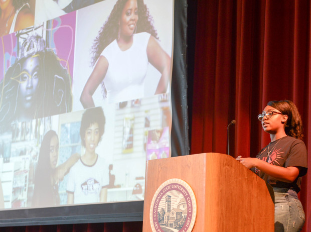 Darlasia Miller speaks from a podium with a powerpoint behind her of black women and their hair.