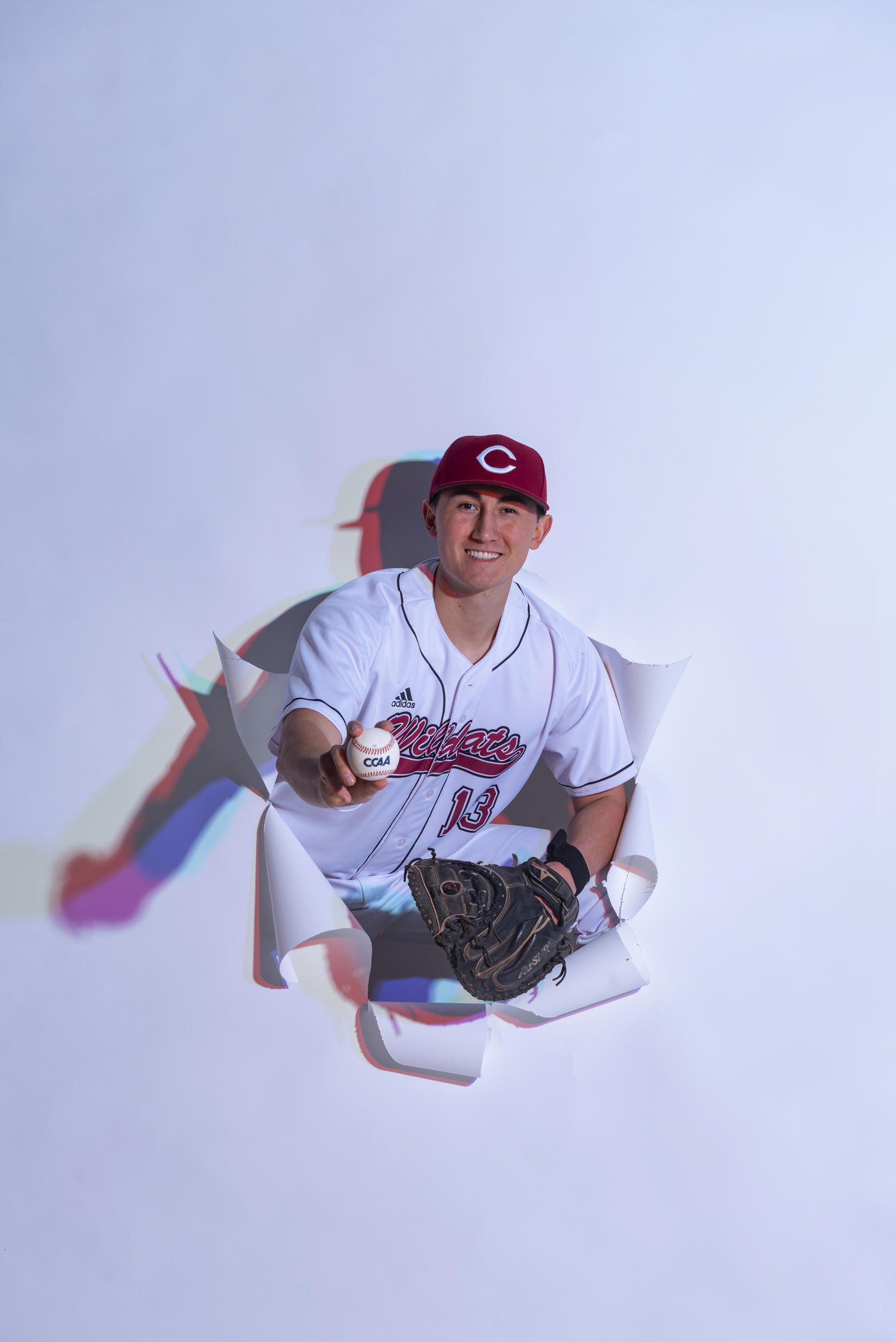 Tyler Stofiel breaks through a white photo backdrop in his uniform and a baseball in hand.