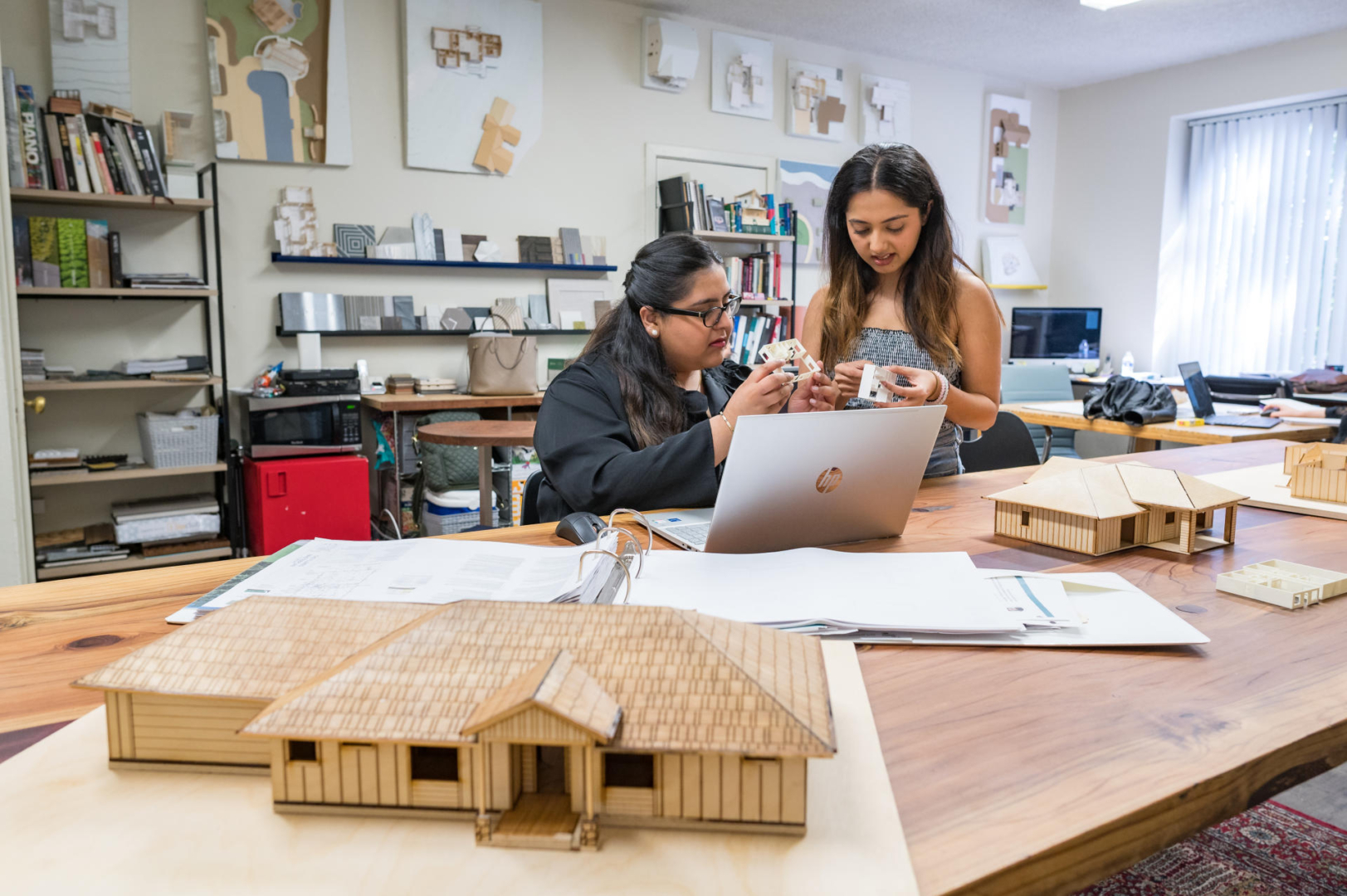 Charvi Grover and Hira Namit hold tiny plastic models of homes in their hands while working on a laptop in the design studio. A wooden model of the house sits on the table in front of them.