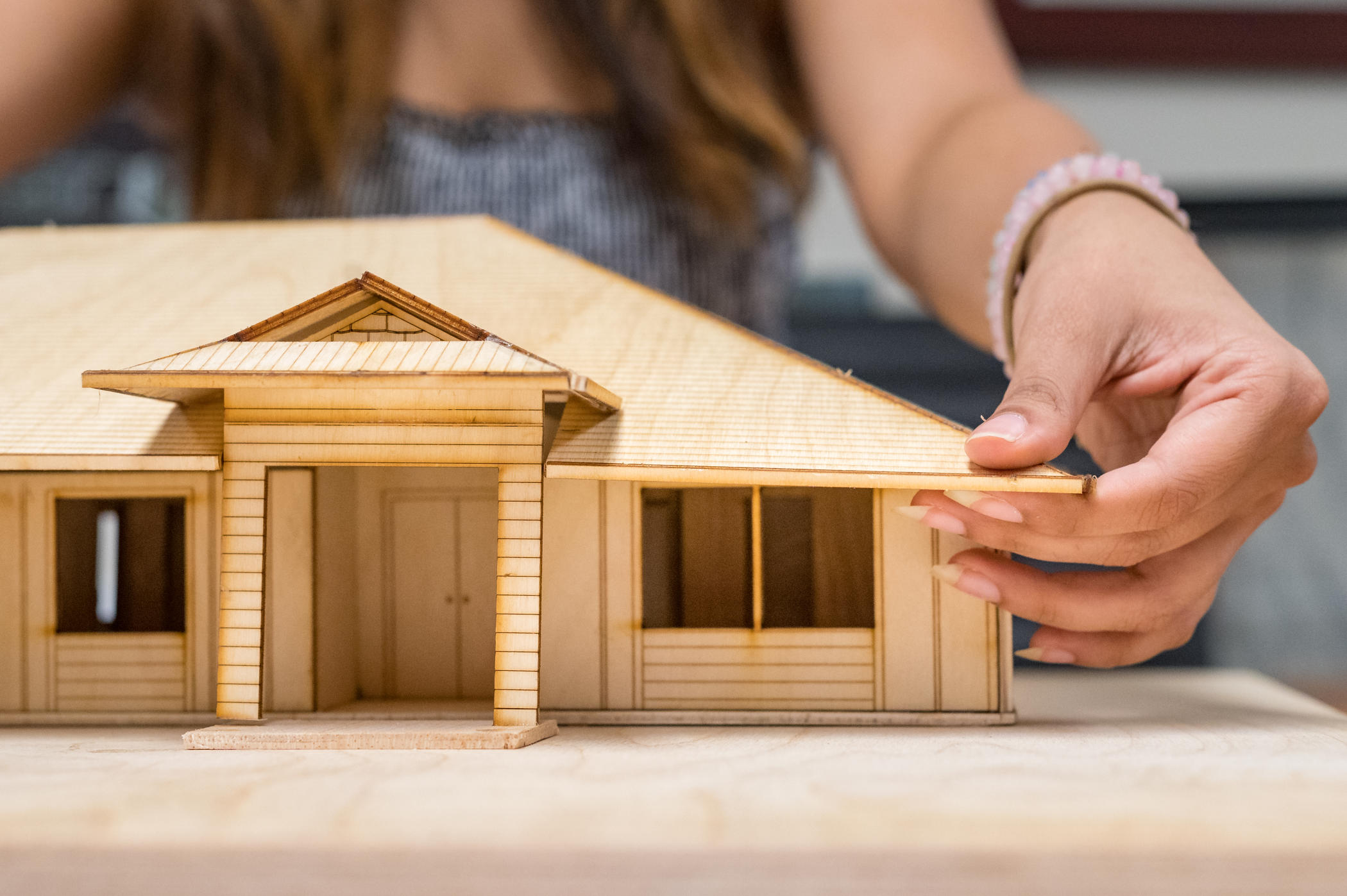Hands adjust the positioning of a roof on a small wooden model of a home, with the view cast toward the front entrance with a covered porch flanked by large windows.