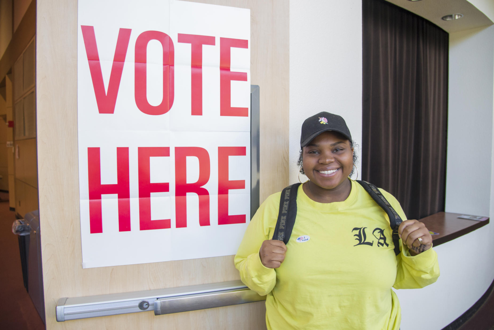 A student stands next to a "vote here" sign while wearing an "I voted" sticker.