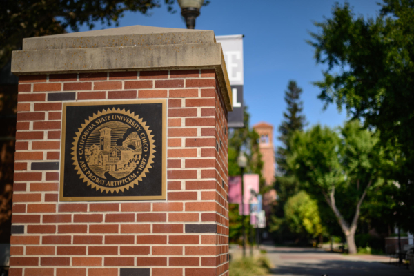 A plaque of Chico State's seal is mounted on a brick pillar, with blue skies and lush trees in the background.
