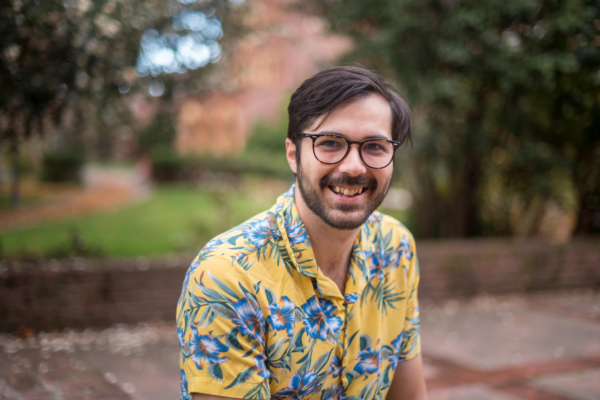 Max Cordeiro, Chico State’s inaugural Human Resources Equity, Diversity, and Inclusion Recruitment and Retention Specialist, poses on the patio outside of Laxson Auditorium. He is smiling joyfully and wearing a yellow Hawaiin-style collared shirt featuring blue and orange flowers. Kendall Hall and lush green plants are in the background.