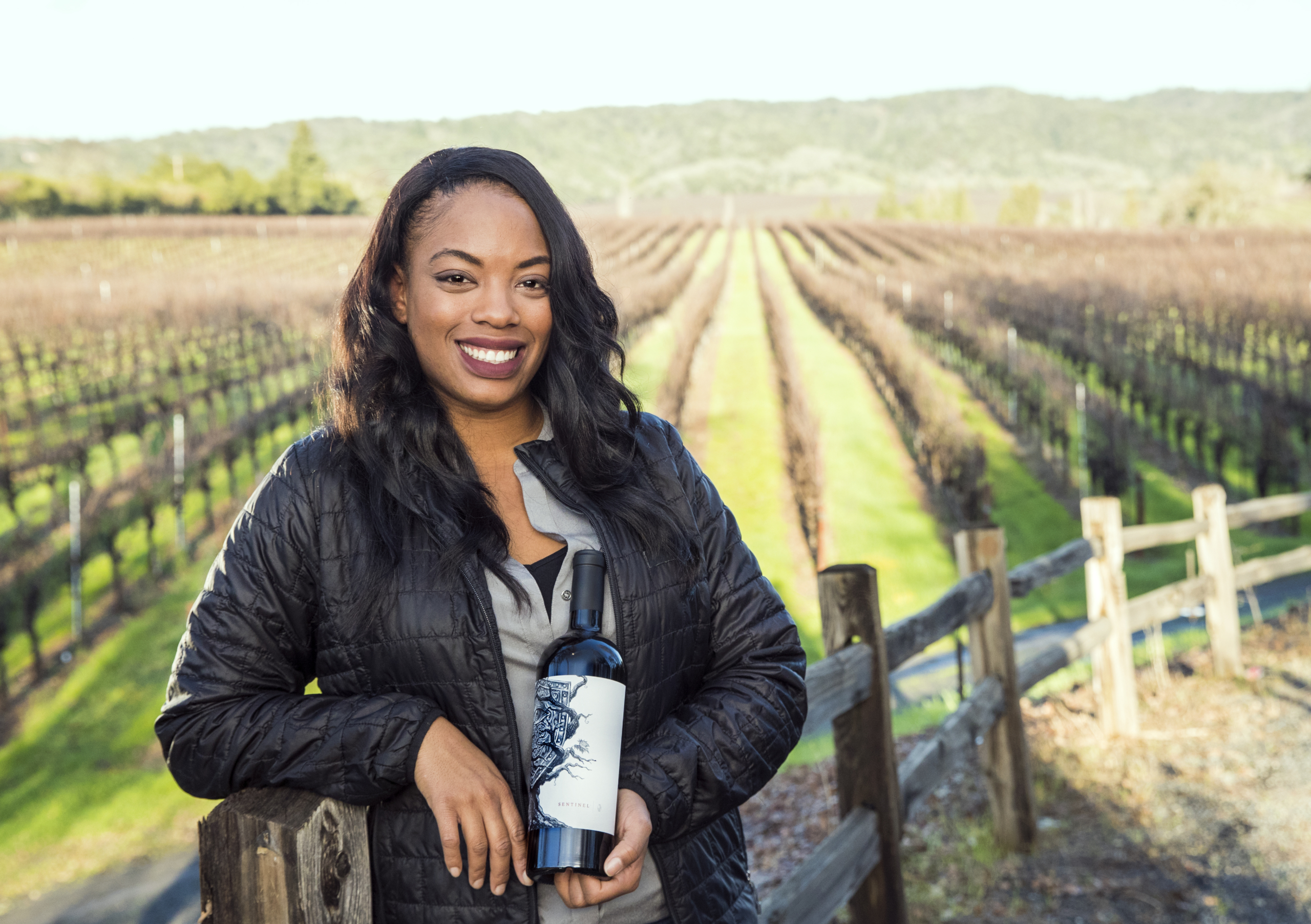 Brenae Royal holds a bottle of wine with a vineyard in the backdrop
